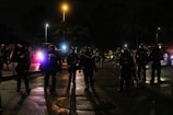Several people in heavy gear and helmets stand in formation on a dimly lit street. They seem to be part of a security or police unit, accompanied by motorcycles in the background. The atmosphere is tense, with lights from vehicles casting shadows on the wet pavement.
