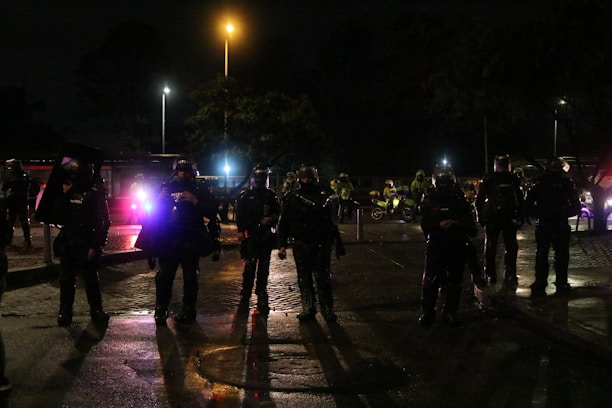 Several people in heavy gear and helmets stand in formation on a dimly lit street. They seem to be part of a security or police unit, accompanied by motorcycles in the background. The atmosphere is tense, with lights from vehicles casting shadows on the wet pavement.