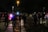 Several people in heavy gear and helmets stand in formation on a dimly lit street. They seem to be part of a security or police unit, accompanied by motorcycles in the background. The atmosphere is tense, with lights from vehicles casting shadows on the wet pavement.
