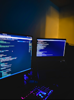 A dimly lit room with two computer monitors displaying code, accompanied by a glowing keyboard in vibrant colors. The monitors show text-based programming interfaces, with one screen filled with lines of code. The surrounding environment is dark, adding to the focus on the screens and keyboard.