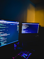 A dimly lit room with two computer monitors displaying code, accompanied by a glowing keyboard in vibrant colors. The monitors show text-based programming interfaces, with one screen filled with lines of code. The surrounding environment is dark, adding to the focus on the screens and keyboard.