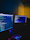 A dimly lit room with two computer monitors displaying code, accompanied by a glowing keyboard in vibrant colors. The monitors show text-based programming interfaces, with one screen filled with lines of code. The surrounding environment is dark, adding to the focus on the screens and keyboard.