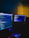 A dimly lit room with two computer monitors displaying code, accompanied by a glowing keyboard in vibrant colors. The monitors show text-based programming interfaces, with one screen filled with lines of code. The surrounding environment is dark, adding to the focus on the screens and keyboard.