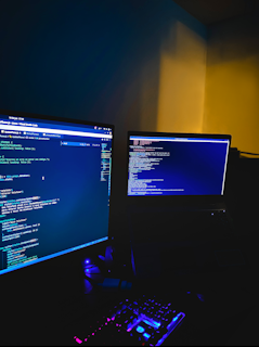 A dimly lit room with two computer monitors displaying code, accompanied by a glowing keyboard in vibrant colors. The monitors show text-based programming interfaces, with one screen filled with lines of code. The surrounding environment is dark, adding to the focus on the screens and keyboard.