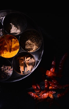 A metal plate contains several bowls filled with various spices, including turmeric powder, cinnamon sticks, cumin, coriander seeds, and cloves. Next to the plate, dried red chili peppers are scattered against a dark background, creating a vibrant contrast.