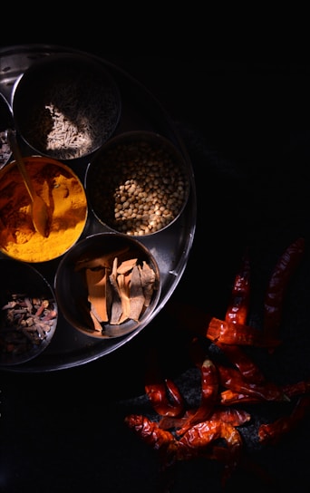 A metal plate contains several bowls filled with various spices, including turmeric powder, cinnamon sticks, cumin, coriander seeds, and cloves. Next to the plate, dried red chili peppers are scattered against a dark background, creating a vibrant contrast.