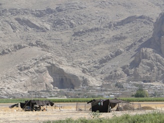A rugged, mountainous landscape with two black tents in the foreground. The background reveals rocky hills and a small settlement with structures that blend into the natural terrain. Surrounding the tents, there are patches of cultivated land, adding contrast to the otherwise barren surroundings.