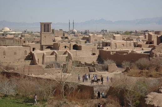 An ancient cityscape with traditional mud-brick buildings and a prominent windcatcher tower against a backdrop of distant mountains. People on horseback and others walking in a dry, rural area in the foreground, surrounded by sparse vegetation.