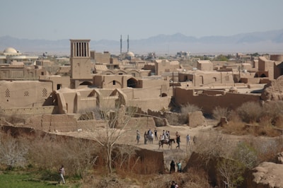 An ancient cityscape with traditional mud-brick buildings and a prominent windcatcher tower against a backdrop of distant mountains. People on horseback and others walking in a dry, rural area in the foreground, surrounded by sparse vegetation.
