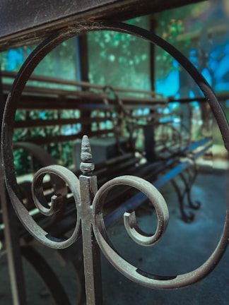 A close-up of ornate metalwork featuring a circular and spiral design with a central pointed tip. The background shows a blurry view of what appears to be additional metal structures, possibly furniture, against a backdrop of greenery and dappled sunlight.