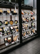 A display window with neatly arranged plastic food models representing various Japanese dishes. The models are placed on shelves, each accompanied by a label with descriptions and prices. The setup is in a traditional Japanese style, featuring wooden frames and a tiled roof.