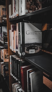 A bookshelf filled with a variety of books positioned closely together. The book 'Giresun' is prominently displayed, sticking out slightly more than others. The lighting is dim, giving the scene a quiet, contemplative atmosphere typical of a library setting. The shelves are made of dark wood, and the overall arrangement appears organized and maintained.