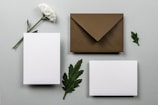 A white flower with a green stem and leaves is placed next to a dark brown envelope. Two blank white cards are positioned nearby on a light grey surface. Green leaves are arranged around the objects, creating a minimalist and elegant composition.