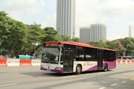 A public transit bus marked 'SBS Transit' is traveling on a city street. The bus is predominantly white and purple with some red accents. In the background, several tall modern buildings with unique architectural designs can be seen, along with lush green trees lining the street. Road barriers in orange and white are positioned alongside the road.