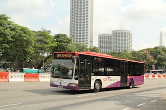 A public transit bus marked 'SBS Transit' is traveling on a city street. The bus is predominantly white and purple with some red accents. In the background, several tall modern buildings with unique architectural designs can be seen, along with lush green trees lining the street. Road barriers in orange and white are positioned alongside the road.