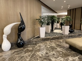A modern indoor setting with a polished marble floor and a decorative area featuring white and black abstract sculptures. Tall white planters with green plants add a natural touch. The background includes a stylish grid-like wall design providing texture and depth.