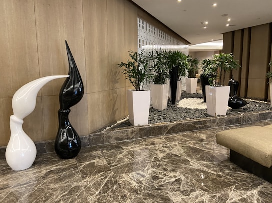 A modern indoor setting with a polished marble floor and a decorative area featuring white and black abstract sculptures. Tall white planters with green plants add a natural touch. The background includes a stylish grid-like wall design providing texture and depth.