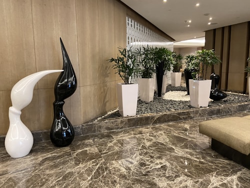 A modern indoor setting with a polished marble floor and a decorative area featuring white and black abstract sculptures. Tall white planters with green plants add a natural touch. The background includes a stylish grid-like wall design providing texture and depth.