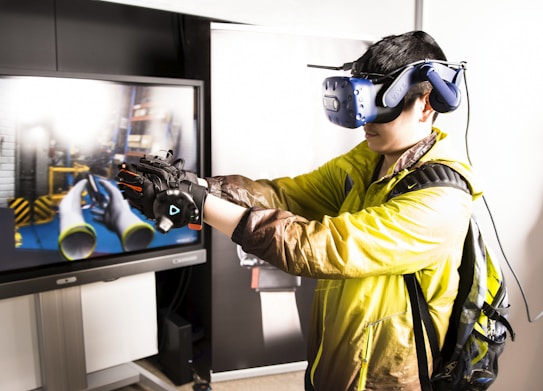 A person is engaged in a virtual reality experience, wearing a VR headset and gloves. They are facing a large screen displaying a simulated environment with tubes and industrial elements. The individual is wearing a yellow jacket and a backpack, appearing focused on the VR interaction.