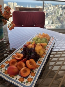 A beautifully arranged platter of fresh fruits sits on a glass table. The platter includes halved apricots, grapes, apple slices, and peaches. The background reveals a scenic cityscape and a view of the sea, adding a sense of spaciousness and luxury. A red cushion and a potted plant with orange flowers add to the aesthetic setup.