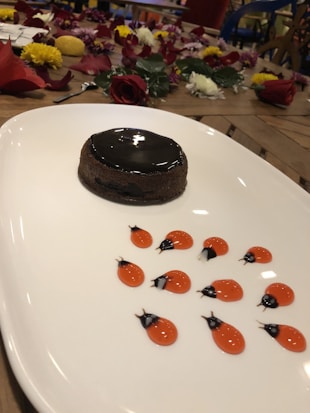 A beautifully plated dessert with a glossy chocolate cake on a white plate. The plate is decorated with artful red and black sauce designs resembling ladybugs. In the background, there is a table adorned with scattered flower petals and whole flowers in various colors.