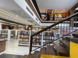 A spacious, modern library with multiple levels and rows of bookshelves. The lower level is filled with neatly organized books, while the upper level has people browsing. The design features a combination of wooden and contemporary elements with bright lighting and minimalist decor.