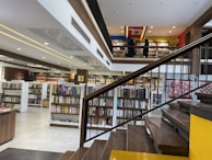 A spacious, modern library with multiple levels and rows of bookshelves. The lower level is filled with neatly organized books, while the upper level has people browsing. The design features a combination of wooden and contemporary elements with bright lighting and minimalist decor.