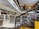 A spacious, modern library with multiple levels and rows of bookshelves. The lower level is filled with neatly organized books, while the upper level has people browsing. The design features a combination of wooden and contemporary elements with bright lighting and minimalist decor.