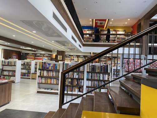 A spacious, modern library with multiple levels and rows of bookshelves. The lower level is filled with neatly organized books, while the upper level has people browsing. The design features a combination of wooden and contemporary elements with bright lighting and minimalist decor.