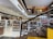A spacious, modern library with multiple levels and rows of bookshelves. The lower level is filled with neatly organized books, while the upper level has people browsing. The design features a combination of wooden and contemporary elements with bright lighting and minimalist decor.