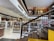 A spacious, modern library with multiple levels and rows of bookshelves. The lower level is filled with neatly organized books, while the upper level has people browsing. The design features a combination of wooden and contemporary elements with bright lighting and minimalist decor.