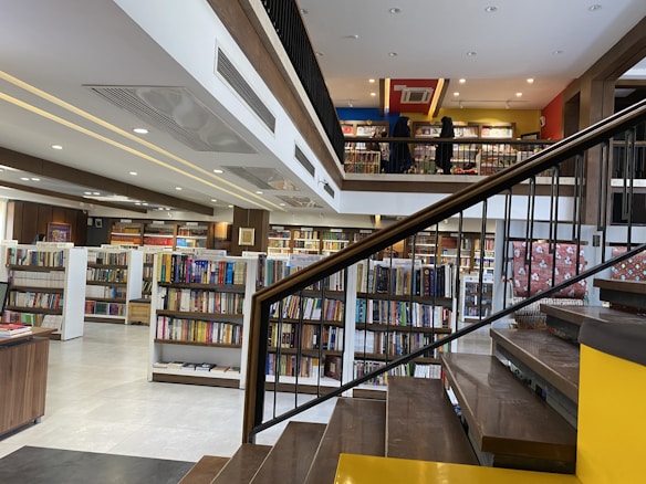 A spacious, modern library with multiple levels and rows of bookshelves. The lower level is filled with neatly organized books, while the upper level has people browsing. The design features a combination of wooden and contemporary elements with bright lighting and minimalist decor.