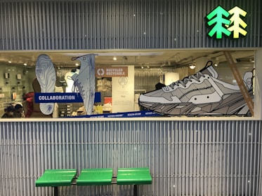 A store display window showcases large, line-art illustrations of shoes with emphasis on a collaboration and recycling theme. The background includes a mix of retail items and people, suggesting a store interior. Above the display is a bright green and yellow icon resembling trees. Below the window, there's a bench with a green seat.