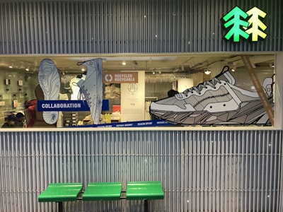 A store display window showcases large, line-art illustrations of shoes with emphasis on a collaboration and recycling theme. The background includes a mix of retail items and people, suggesting a store interior. Above the display is a bright green and yellow icon resembling trees. Below the window, there's a bench with a green seat.