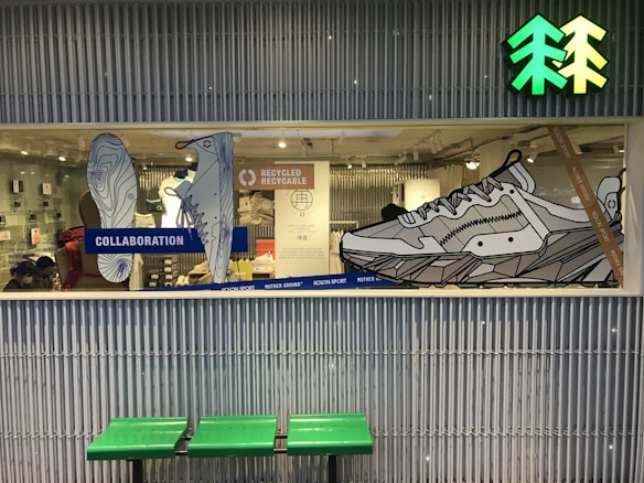 A store display window showcases large, line-art illustrations of shoes with emphasis on a collaboration and recycling theme. The background includes a mix of retail items and people, suggesting a store interior. Above the display is a bright green and yellow icon resembling trees. Below the window, there's a bench with a green seat.