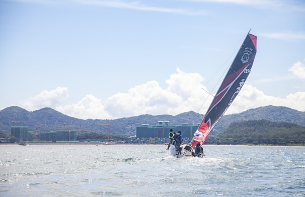 A sailing boat with a large black and red sail glides through the water, with a crew on board. The background features distant mountains and green-covered buildings along the shoreline. The sky is partly cloudy, suggesting a bright, fair-weather day.