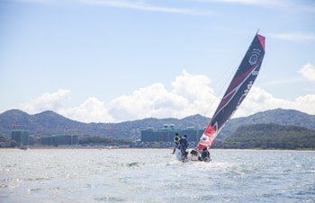 A sailing boat with a large black and red sail glides through the water, with a crew on board. The background features distant mountains and green-covered buildings along the shoreline. The sky is partly cloudy, suggesting a bright, fair-weather day.