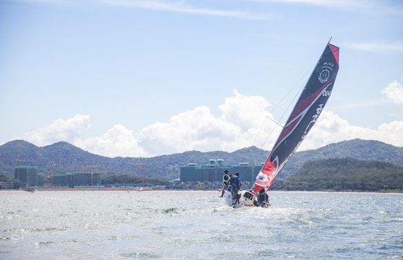 A sailing boat with a large black and red sail glides through the water, with a crew on board. The background features distant mountains and green-covered buildings along the shoreline. The sky is partly cloudy, suggesting a bright, fair-weather day.