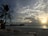 A tranquil beach scene at sunset showcases a vast sky filled with dramatic clouds. A single palm tree stands to the left, and in the background, overwater bungalows stretch out into the ocean. The foreground includes several wooden tables and chairs on the sandy shore.