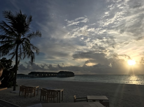A tranquil beach scene at sunset showcases a vast sky filled with dramatic clouds. A single palm tree stands to the left, and in the background, overwater bungalows stretch out into the ocean. The foreground includes several wooden tables and chairs on the sandy shore.
