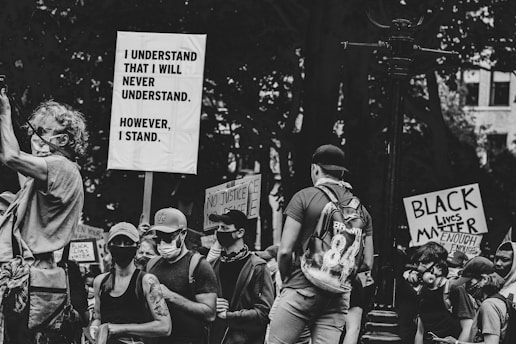 A crowd of people, many wearing masks, hold various signs that express messages related to social justice and solidarity. The image is in black and white, adding a serious, poignant effect to the scene.