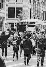 A group of people are marching down the street, holding signs with social justice messages. They are walking past a bus with the destination 'Grand St' displayed. The participants appear united and purposeful as they hold their fists in the air.