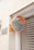 A convex mirror with an orange frame is mounted on a brick wall next to a sign with Japanese characters. The reflection in the mirror shows a distorted view of the gridded metal window shutter opposite it. The wall is made of light beige bricks, and there is an orange hue from the rust-colored panel that holds the mirror.