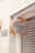 A convex mirror with an orange frame is mounted on a brick wall next to a sign with Japanese characters. The reflection in the mirror shows a distorted view of the gridded metal window shutter opposite it. The wall is made of light beige bricks, and there is an orange hue from the rust-colored panel that holds the mirror.