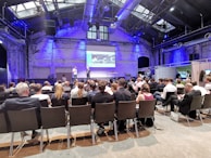 A conference room filled with attendees seated and facing a stage where two speakers stand in front of a presentation screen displaying an image. The room has high ceilings with industrial architecture and blue lighting.