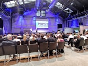 A conference room filled with attendees seated and facing a stage where two speakers stand in front of a presentation screen displaying an image. The room has high ceilings with industrial architecture and blue lighting.