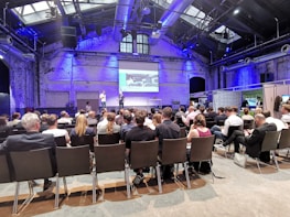 A conference room filled with attendees seated and facing a stage where two speakers stand in front of a presentation screen displaying an image. The room has high ceilings with industrial architecture and blue lighting.