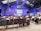 A conference room filled with attendees seated and facing a stage where two speakers stand in front of a presentation screen displaying an image. The room has high ceilings with industrial architecture and blue lighting.