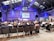 A conference room filled with attendees seated and facing a stage where two speakers stand in front of a presentation screen displaying an image. The room has high ceilings with industrial architecture and blue lighting.