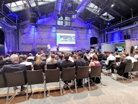 A conference room filled with attendees seated and facing a stage where two speakers stand in front of a presentation screen displaying an image. The room has high ceilings with industrial architecture and blue lighting.